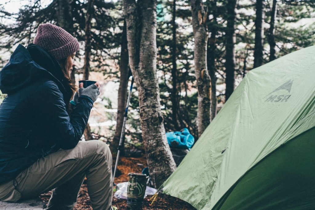 A woman enjoying a cup of coffee outside of her tent in a forest setting. She packed the essential coping gear for her outdoor adventure.