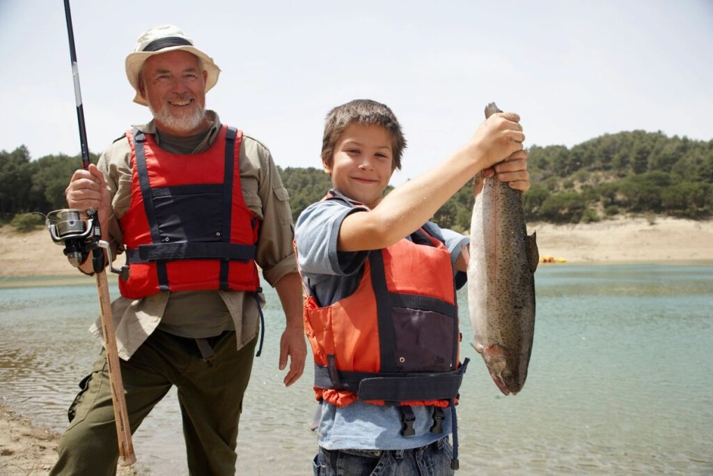 Grandfather and grandson on a fishing trip and the grandson is holding a very large tout they he just got. The grandfather is very happy.